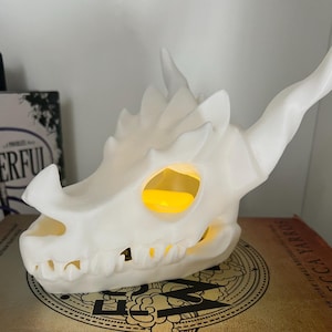 May include: White 3D printed dragon skull candle holder with a glowing yellow light inside. The candle holder is sitting on top of two books, one with the title "Fourth Wing" and the other with the title "Iron Flame".