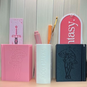 Puede incluir: Tres sujetalibros en rosa, blanco y negro con un diseño de hongo y luna. Los sujetalibros están decorados con las palabras "Bookmarks" y "Fantasy".
