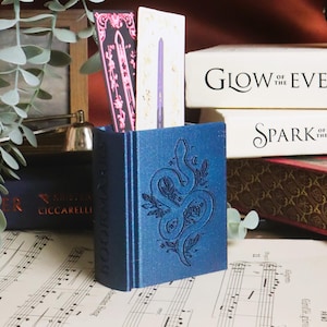 Könnte beinhalten: Ein blauer, buchförmiger Lesezeichenhalter mit einem Schlangen- und Blumendesign. Zwei Lesezeichen sind im Halter sichtbar. Der Halter steht auf einer Oberfläche mit Noten. Weitere Bücher sind im Hintergrund.