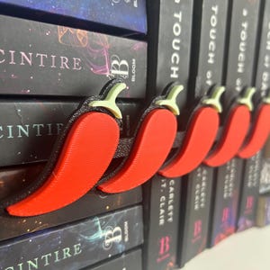Puede incluir: Un conjunto de cinco sujetap&aacute;ginas con forma de pimiento rojo. Cada pimiento tiene un tallo verde y un contorno negro. Los accesorios para libros se muestran frente a una pila de libros.