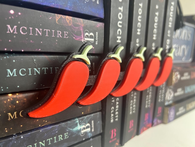 Puede incluir: Un conjunto de cinco sujetap&aacute;ginas con forma de pimiento rojo. Cada pimiento tiene un tallo verde y un contorno negro. Los accesorios para libros se muestran frente a una pila de libros.