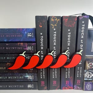 Puede incluir: Una fila de marcadores de pimiento rojo con tallos blancos y bordes negros se coloca encima de una pila de libros. Los libros tienen lomos negros con el t&iacute;tulo "a TOUCH of..." y nombres de autores.