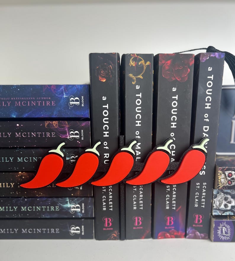Puede incluir: Una fila de marcadores de pimiento rojo con tallos blancos y bordes negros se coloca encima de una pila de libros. Los libros tienen lomos negros con el t&iacute;tulo "a TOUCH of..." y nombres de autores.