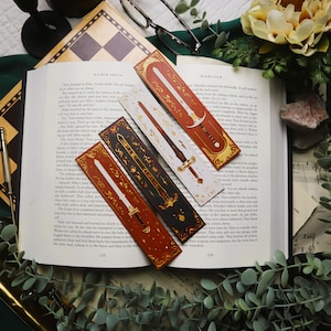 May include: Four decorative bookmarks with sword designs in shades of red, black, and white, with gold accents. The bookmarks are rectangular and feature detailed illustrations of swords. They are placed on an open book.