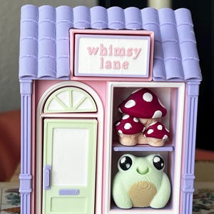 May include: A miniature shop structure with a lavender roof and pink walls. A sign above the door reads "whimsy lane". The shop features a light green door and a display window containing a cluster of red mushrooms and a light green frog figurine.