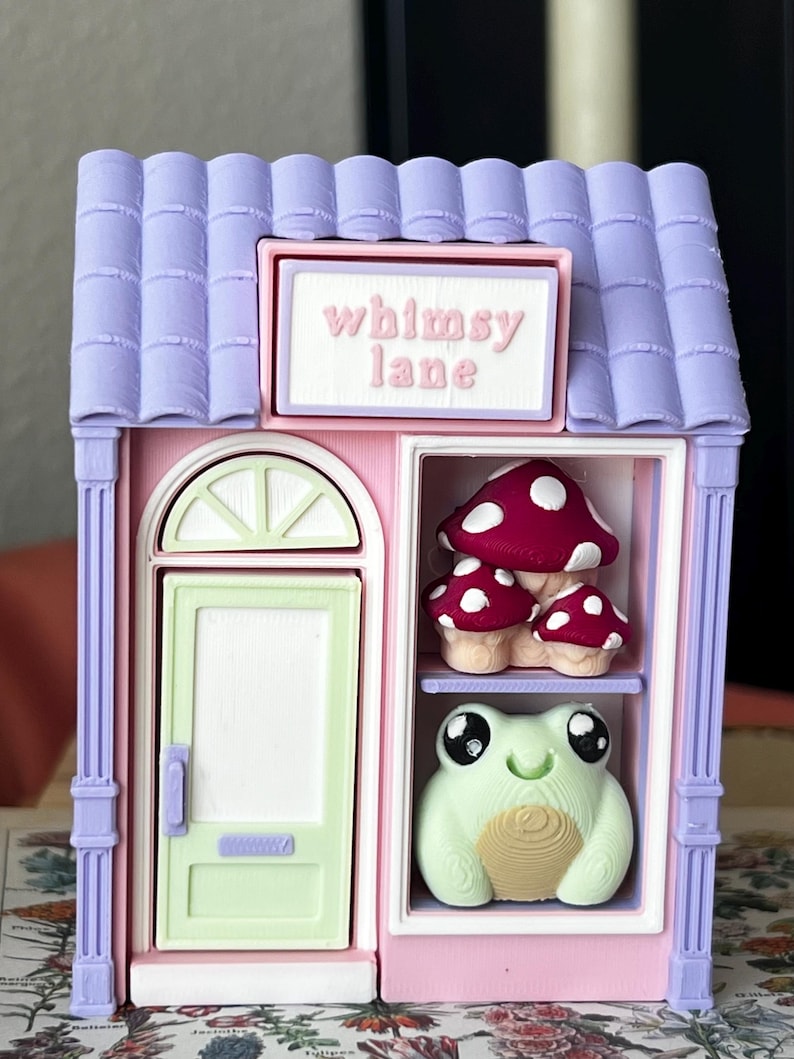 May include: A miniature shop structure with a lavender roof and pink walls. A sign above the door reads "whimsy lane". The shop features a light green door and a display window containing a cluster of red mushrooms and a light green frog figurine.