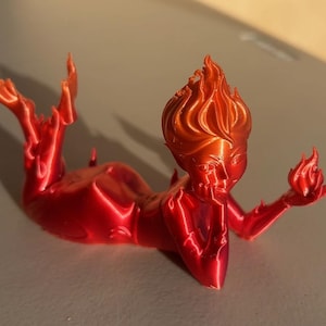 May include: A red and orange figurine of a reclining figure with flames for hair and holding a flame in their hand. The figurine is a stylized representation of a fire elemental.