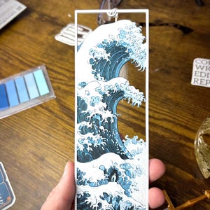 May include: A rectangular bookmark featuring a stylized depiction of a large ocean wave in shades of blue and white. The wave design is set within a white rectangular frame. The bookmark is held by a hand.