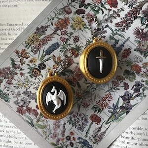May include: Two oval-shaped pendants with gold-toned frames and black centers. One pendant features a white dragon relief, while the other displays a silver-toned dagger. Both items include a small metal ball chain for attachment or hanging.
