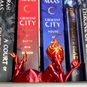 May include: A red and orange 3D-printed bookmark in the shape of a reclining figure holding a flame. The bookmark is placed among a row of books with titles including "Crescent City" and "Throne of Glass".