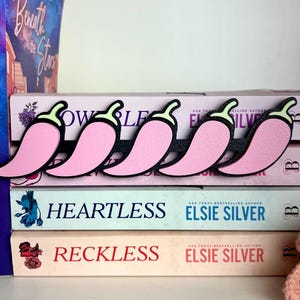 Puede incluir: Marcadores de libros con forma de pimiento rosa con contornos negros y tallos verdes colocados sobre una pila de libros. Los libros tienen t&iacute;tulos como "Heartless" y "Reckless" de Elsie Silver. Se ven lomos de otros libros en el fondo.