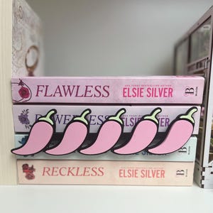 Puede incluir: Sujetalibros rosas con forma de chile y contornos negros, colocados sobre una pila de libros. Los libros se titulan "Flawless", "Reckless" y "Wees" de Elsie Silver. Los libros son rosas y blancos.