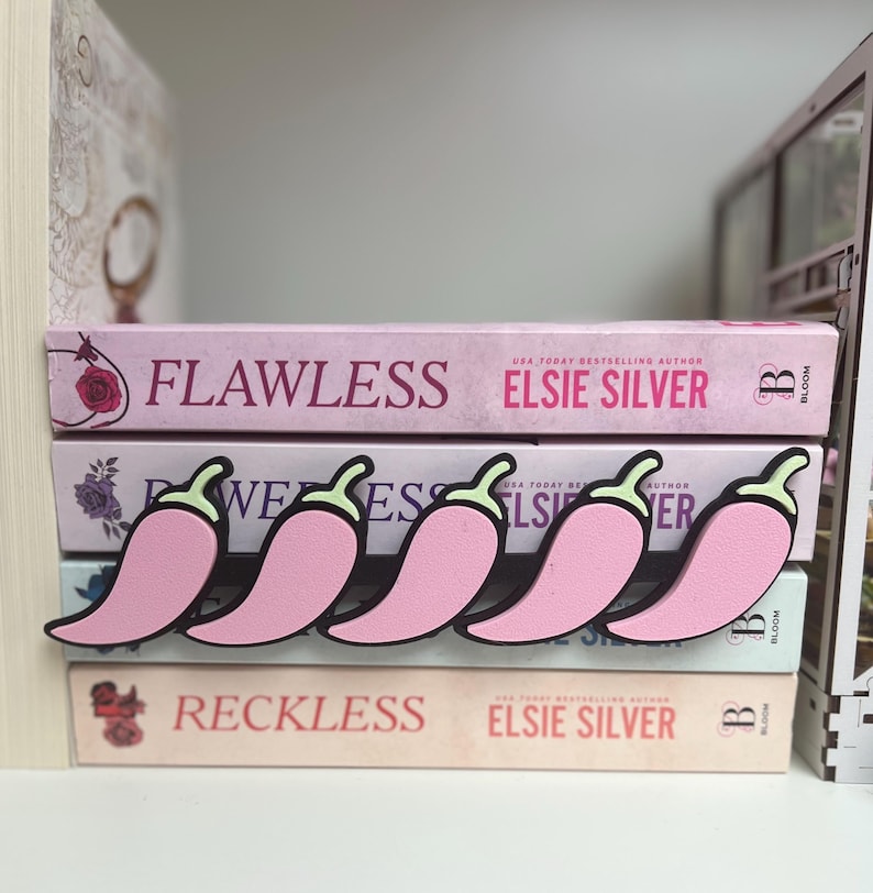 Puede incluir: Sujetalibros rosas con forma de chile y contornos negros, colocados sobre una pila de libros. Los libros se titulan "Flawless", "Reckless" y "Wees" de Elsie Silver. Los libros son rosas y blancos.