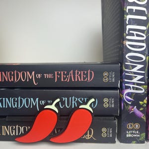 Puede incluir: Dos marcadores con forma de pimiento rojo descansan sobre una pila de libros. Los libros tienen cubiertas negras con t&iacute;tulos en letras blancas y rosas. El libro de la derecha tiene un dise&ntilde;o floral y el t&iacute;tulo "BELLADONNA".