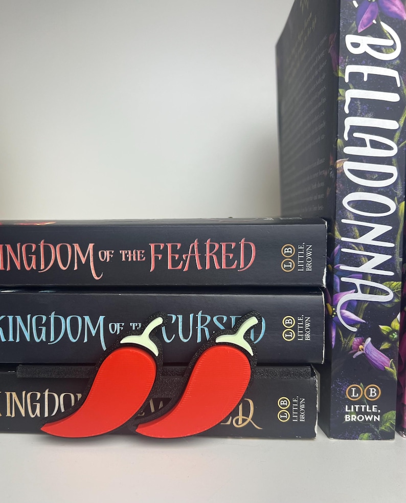 Puede incluir: Dos marcadores con forma de pimiento rojo descansan sobre una pila de libros. Los libros tienen cubiertas negras con t&iacute;tulos en letras blancas y rosas. El libro de la derecha tiene un dise&ntilde;o floral y el t&iacute;tulo "BELLADONNA".