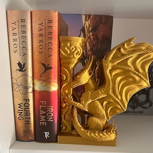 Op de afbeelding: Een goudkleurige drakenboekensteun met gedetailleerde vleugels, geplaatst tussen twee boeken. De boeken heten "Fourth Wing" en "Iron Flame" van Rebecca Yarros.