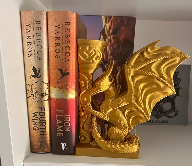 Op de afbeelding: Een goudkleurige drakenboekensteun met gedetailleerde vleugels, geplaatst tussen twee boeken. De boeken heten "Fourth Wing" en "Iron Flame" van Rebecca Yarros.
