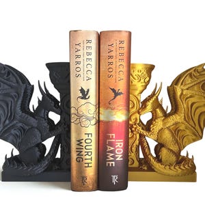 Op de afbeelding: Draakvormige boekensteunen in zwart en goud. De boekensteunen tonen gedetailleerde drakenbeelden met uitgespreide vleugels. Twee boeken, "Fourth Wing" en "Iron Flame" van Rebecca Yarros, staan tussen de boekensteunen. De boeken hebben gouden en oranje covers.