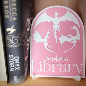 May include: A pink, arched bookend with a white silhouette of three dragons. The bookend has the text "Jordon's Library" in white lettering.