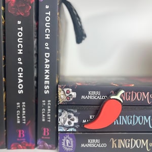 Puede incluir: Un marcap&aacute;ginas rojo con forma de chile descansa sobre una pila de libros. Los libros tienen cubiertas negras con texto blanco e ilustraciones de calaveras. Los lomos de los libros en el estante tienen los t&iacute;tulos "A Touch of Chaos" y "A Touch of Darkness".