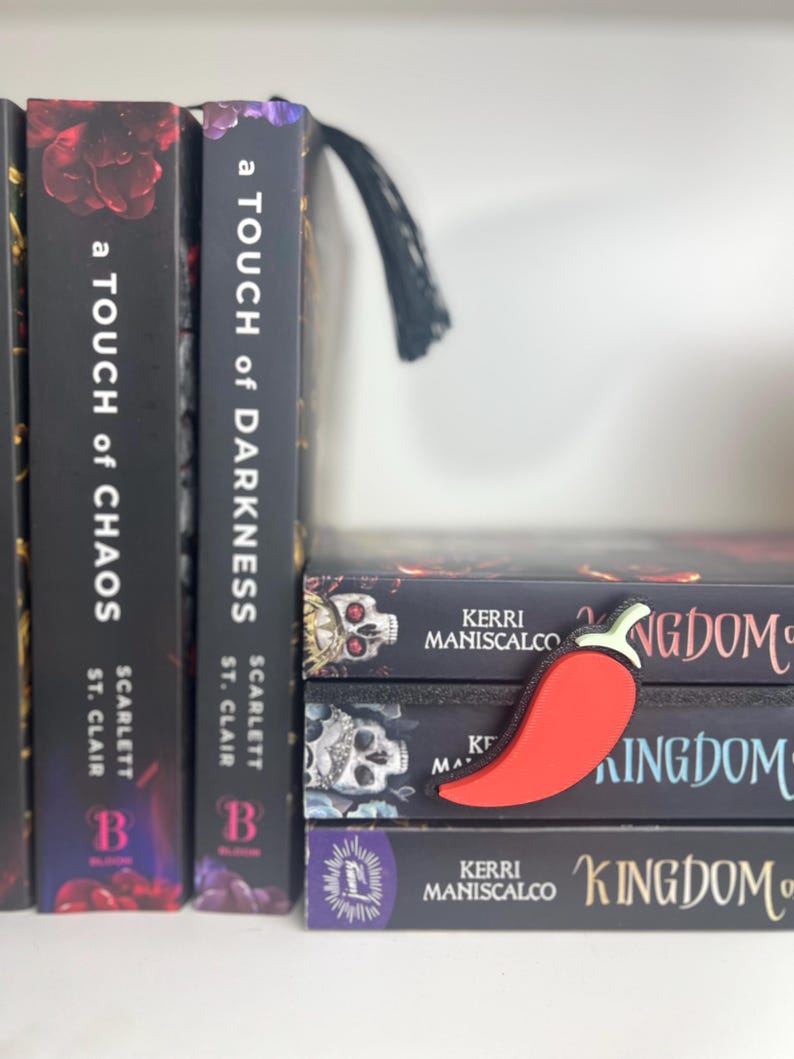 Puede incluir: Un marcap&aacute;ginas rojo con forma de chile descansa sobre una pila de libros. Los libros tienen cubiertas negras con texto blanco e ilustraciones de calaveras. Los lomos de los libros en el estante tienen los t&iacute;tulos "A Touch of Chaos" y "A Touch of Darkness".