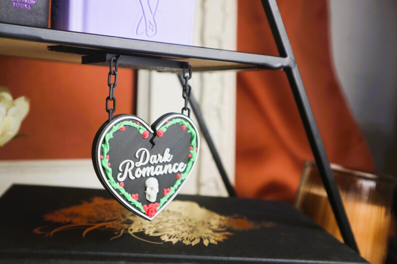 May include: A black heart-shaped sign with the words "Dark Romance" in white script, decorated with red roses, green leaves, and a small skull. The sign hangs from a black chain attached to a black metal shelf.
