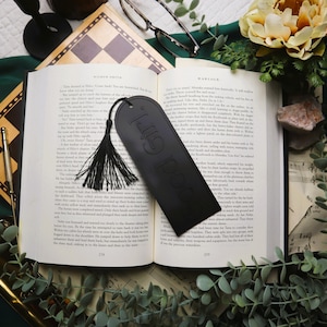 Puede incluir: Un marcapáginas de cuero negro con las palabras "Good Girl" en relieve, colocado en un libro abierto. El marcapáginas tiene una borla negra y está rodeado de objetos decorativos.