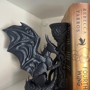 Op de afbeelding: Een zwarte drakenboekensteun met gedetailleerde vleugels en schubben, die een decoratieve pilaar vasthoudt. De draak staat naast een boek met de titel "Fourth Wing" van Rebecca Yarros. De boekensteun staat op een witte plank.