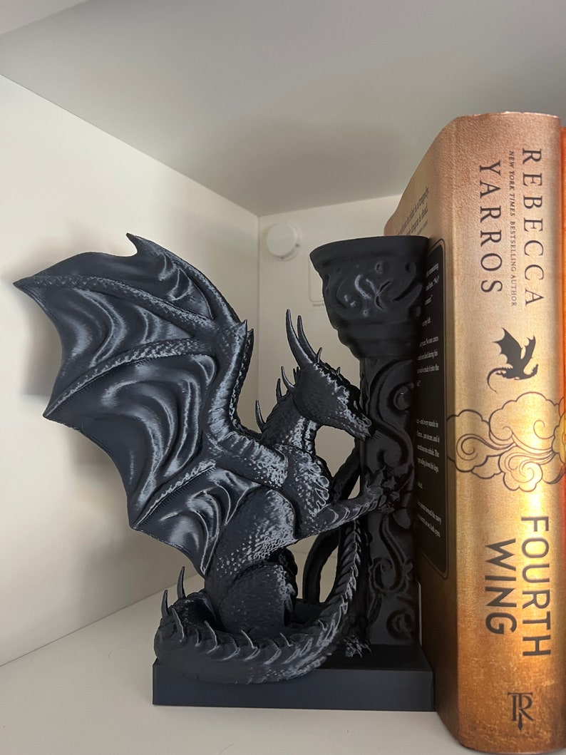 Op de afbeelding: Een zwarte drakenboekensteun met gedetailleerde vleugels en schubben, die een decoratieve pilaar vasthoudt. De draak staat naast een boek met de titel "Fourth Wing" van Rebecca Yarros. De boekensteun staat op een witte plank.