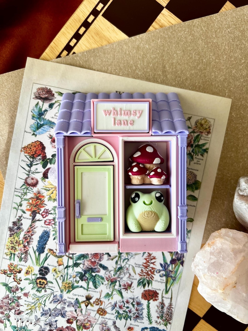 May include: A small, three-dimensional shop structure with a lavender roof and pink walls. A sign above the door reads "whimsy lane" in pink text. The shop features a light green door and a display window containing three red mushrooms and a light green frog.