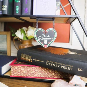 May include: A heart-shaped sign reading "Dark Romance" hangs above a stack of books. The books are black and red, with gold lettering. A vase of white flowers and greenery are also visible.