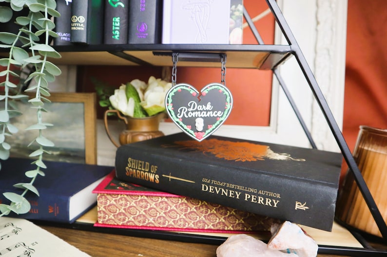 May include: A heart-shaped sign reading "Dark Romance" hangs above a stack of books. The books are black and red, with gold lettering. A vase of white flowers and greenery are also visible.