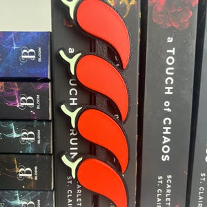 Puede incluir: Un marcap&aacute;ginas de pimiento rojo con una columna vertebral negra, colocado en el lomo de un libro. El marcap&aacute;ginas tiene cuatro pimientos con tallos verdes. Las lomos de los libros son negros con texto blanco.