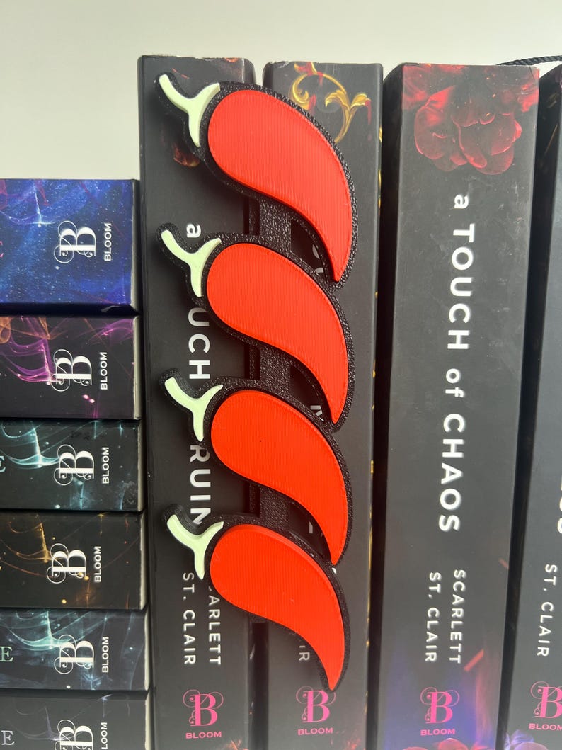 Puede incluir: Un marcap&aacute;ginas de pimiento rojo con una columna vertebral negra, colocado en el lomo de un libro. El marcap&aacute;ginas tiene cuatro pimientos con tallos verdes. Las lomos de los libros son negros con texto blanco.