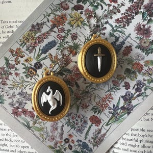 May include: Two oval-shaped pendants with gold-colored frames and black centers. One pendant features a white dragon relief, and the other features a silver-colored sword relief. Each pendant includes a small metal ball chain for attachment.