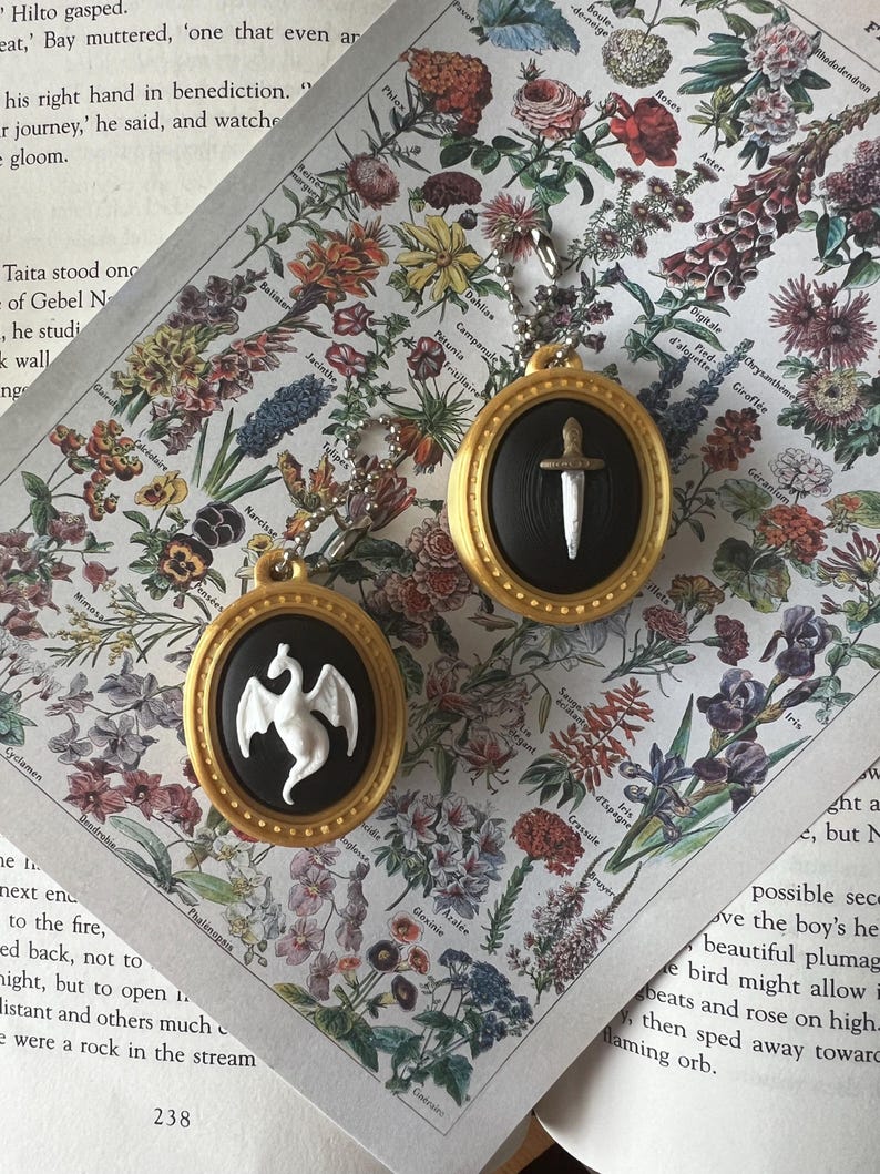 May include: Two oval-shaped pendants with gold-colored frames and black centers. One pendant features a white dragon relief, and the other features a silver-colored sword relief. Each pendant includes a small metal ball chain for attachment.