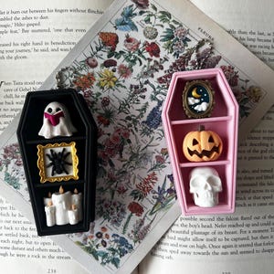 May include: Two coffin-shaped display cases with shelves. The black case contains a ghost reading a book, a spider in a gold frame, and white candles. The pink case holds a moon and clouds in a frame, a jack-o'-lantern, and a white skull.