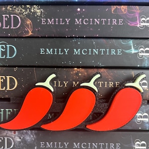 Puede incluir: Tres marcap&aacute;ginas de pimiento rojo con contornos negros y tallos verdes descansan sobre tres libros. Los libros tienen el t&iacute;tulo "ED" y el nombre del autor, "EMILY MCINTIRE", impresos en el lomo.