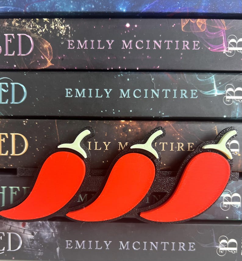 Puede incluir: Tres marcap&aacute;ginas de pimiento rojo con contornos negros y tallos verdes descansan sobre tres libros. Los libros tienen el t&iacute;tulo "ED" y el nombre del autor, "EMILY MCINTIRE", impresos en el lomo.