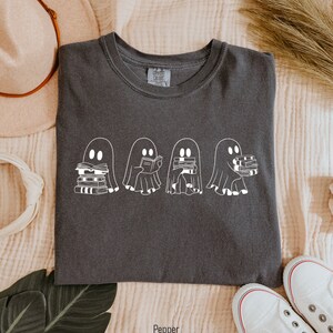May include: A dark gray t-shirt with a white graphic of four ghosts reading books. The ghosts are lined up in a row and each ghost is holding a stack of books. The text "Pepper" is printed below the image.