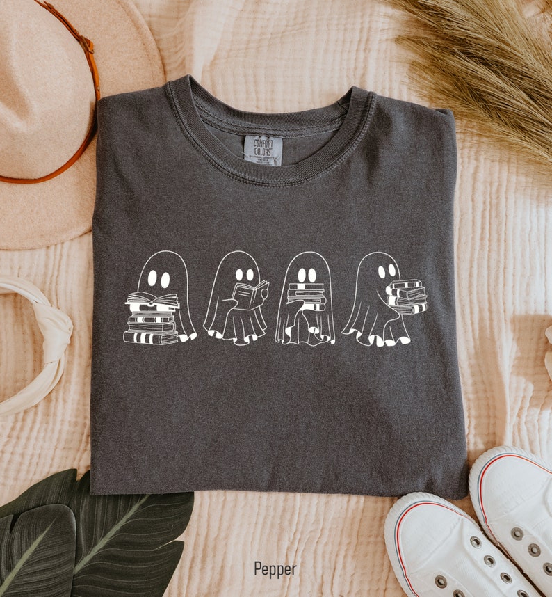 May include: A dark gray t-shirt with a white graphic of four ghosts reading books. The ghosts are lined up in a row and each ghost is holding a stack of books. The text "Pepper" is printed below the image.