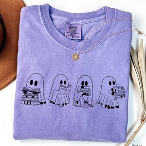 May include: A purple t-shirt with a graphic of four ghosts holding stacks of books. The text "Comfort Colors" is on the tag.