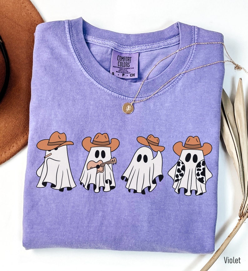 May include: Purple t-shirt with a graphic of four ghosts wearing cowboy hats. One ghost is playing a guitar. The text "Comfort Colors" is on the tag.