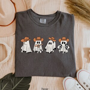 May include: A dark gray t-shirt with a graphic of four ghosts wearing cowboy hats. The ghosts are playing musical instruments and holding a cow print. The text "Pepper" is printed below the image.