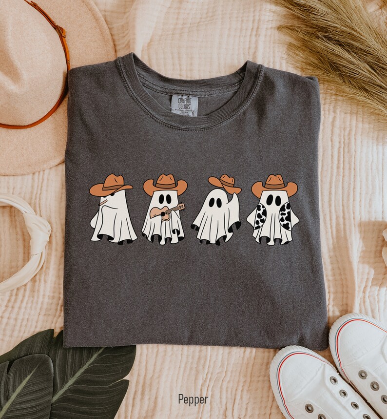 May include: A dark gray t-shirt with a graphic of four ghosts wearing cowboy hats. The ghosts are playing musical instruments and holding a cow print. The text "Pepper" is printed below the image.