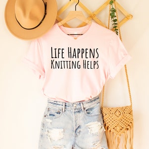 May include: A soft pink t-shirt with the text "Life Happens Knitting Helps" in black lettering. The shirt is hanging on a wooden hanger and is paired with denim shorts and a straw bag.