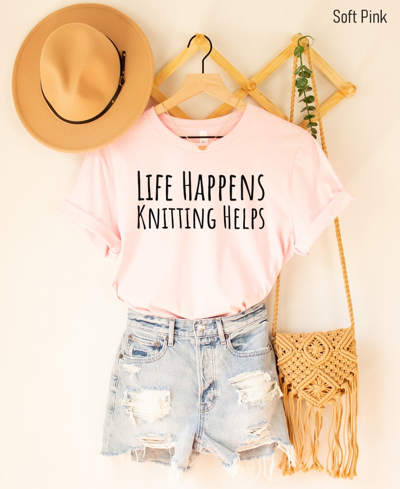 May include: A soft pink t-shirt with the text "Life Happens Knitting Helps" in black lettering. The shirt is hanging on a wooden hanger and is paired with denim shorts and a straw bag.
