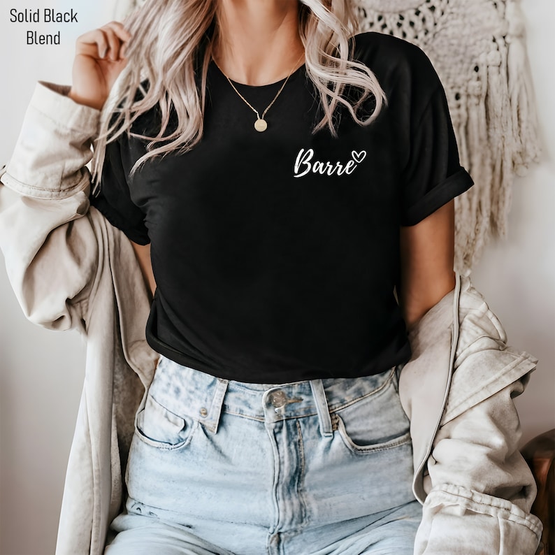 May include: A black t-shirt with a white graphic that says "Barre" with a heart symbol. The shirt is being worn by a person with long blonde hair and is paired with blue jeans.