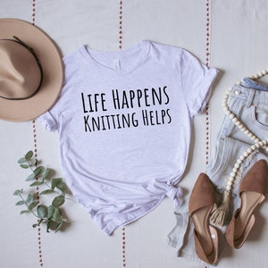 May include: A white t-shirt with the text "Life Happens Knitting Helps" in black lettering. The shirt is laid out on a white and brown patterned surface with a brown hat, a pair of brown shoes, and a pair of blue jeans with a white beaded necklace.