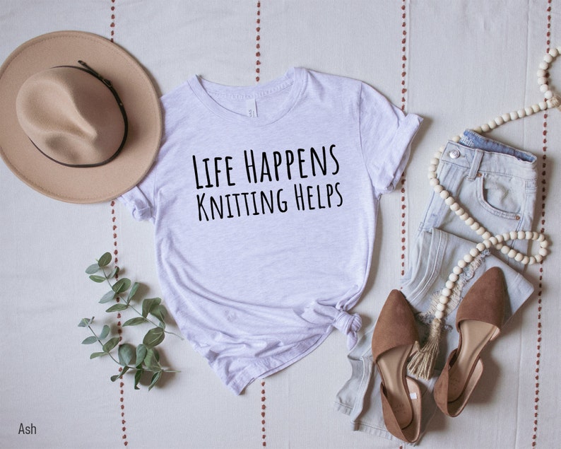 May include: A white t-shirt with the text "Life Happens Knitting Helps" in black lettering. The shirt is laid out on a white and brown patterned surface with a brown hat, a pair of brown shoes, and a pair of blue jeans with a white beaded necklace.